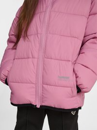 Personne portant une veste matelassée rose zippée avec les mains dans les poches, associée à un pantalon noir avec un petit logo près du genou.
