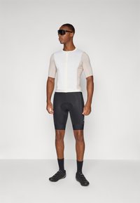 Kortærmet cykeltrøje i hvid og lys beige med lynlås foran, parret med sorte polstrede shorts og sorte cykelsko.