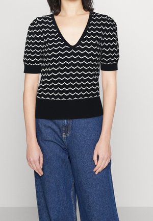 Kobieta w czarnym swetrze z krótkim rękawem i dekoltem w serek, ozdobionym białym wzorem w zygzaki, w połączeniu z niebieskimi, szerokimi spodniami z wysokim stanem.