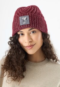 Burgundy knitted beanie with a textured pattern and a gray patch featuring a Snoopy graphic, worn over curly hair, paired with a beige sweater.
