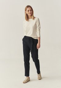 Tricou lung crem deschis, asortat cu pantaloni blugi închiși la culoare, cu curea legată. Încălțămintea constă în adidași bej deschis, cu tălpi texturate.