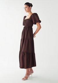 Robe maxi marron avec buste smocké, manches bouffantes et jupe à volants. Associée à des talons ouverts blancs. Texture lisse et silhouette ample.