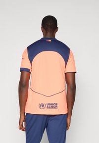 Oranžinis ir tamsiai mėlynas sportinis marškinėlis su apvalia iškirpte, trumpomis rankovėmis, logotipu ant nugaros ir lygiu, kvėpuojančiu audinio paviršiumi.