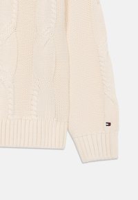 Lichtbeige kabelgebreide trui met geribde manchetten en onderrand, voorzien van een klein logo patch in rood, wit en blauw op de mouw.