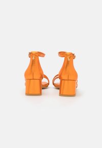 Narancssárga, blokk sarkú szandál, fényes felülettel, boka pántokkal és nyitott orral. Arany részletekkel és textúrált talppal rendelkezik.