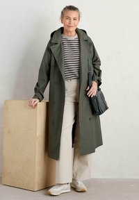 Olijfgroen trenchcoat met grote knopen, gecombineerd met een gestreepte trui en crèmekleurige broek. Accessoires zijn een donkere schoudertas en sneakers.