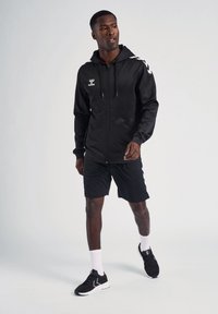 Felpa nera con zip e logo bianco, dettagli sulle spalle, abbinata a pantaloni corti neri, calze bianche e sneakers nere su uno sfondo chiaro.