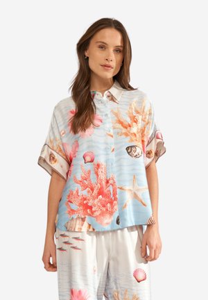 Žena nosi voľnú svetlomodrú a bielu košeľu a nohavice s potlačou koralov, mušlí, hviezdic a rýb, stojaca na bielom pozadí.