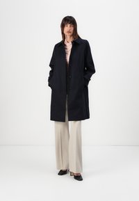 Marineblauer Mantel mit geradem Schnitt, Knopfverschluss und seitlichen Taschen. Trägt man über einem hellrosa Oberteil und kombiniert mit beigen weitgeschnittenen Hosen und schwarzen High Heels.