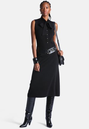Femme portant une robe noire sans manches avec col nœud, large ceinture, bracelet manchette noir, bottes texturées montantes et sac à bandoulière, debout avec confiance.