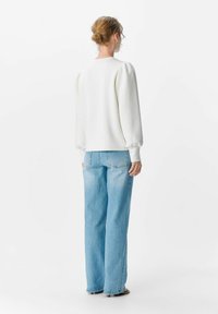 Weißer, strukturierter Pullover mit gepufften Schultern, kombiniert mit hellblauen, weit geschnittenen Jeans. Das Model ist von hinten zu sehen.