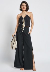 Top corto nero con scollo a halter in tessuto testurizzato e dettagli in pizzo beige, abbinato a pantaloni neri a gamba larga con spacco laterale e bottoni.