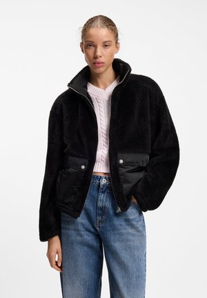 Jeune femme portant une veste noire duveteuse zippée sur un pull en maille torsadée rose clair et un jean bleu, debout devant un fond blanc uni.