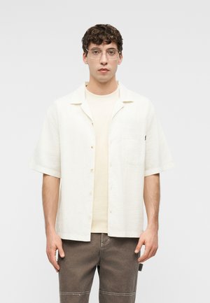 Mladi moški z kodrastimi lasmi, oblečen v bež majico, odprto belo srajco s kratkimi rokavi in rjave hlače, stoji pred enostavnim ozadjem.