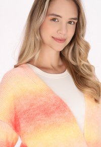 Stickad cardigan i gradientfärger som går från mjuk rosa till gul. Har en v-ringad design och en lätt textur, lagd över en vit topp.