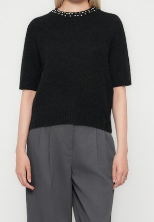 Pull en maille noir à manches courtes avec de petites perles autour du col, porté avec un pantalon plissé gris taille haute.