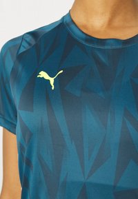 Närbild av en blå sporttröja med geometriskt mönster och en neon-gul Puma-logotyp på vänster bröst, bärs av en person.