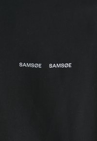 Juodas audinys su dviem baltomis siuvinėtomis „SAM SøE“ logotipais, artimai išdėstytais priekyje, su briaunotu paviršiumi.