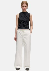 Zwarte mouwloze top met zijruche, gecombineerd met loszittende witte broek met trekkoord en zwarte instappers. Glad stoftextuur zichtbaar.