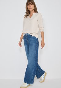 Maglietta beige a maniche lunghe con colletto; jeans blu a gamba larga; sneakers bianche con dettagli blu. Sfondo semplice, modella in piedi.