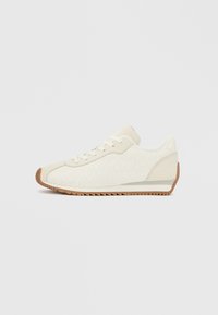 Λευκό υφασμάτινο αθλητικό παπούτσι με εξωτερική σόλα από καουτσούκ καφέ χρώματος, με χαμηλό προφίλ, στρογγυλή μύτη και κλείσιμο με κορδόνια. Λεπτομέρειες στο πλάι περιλαμβάνουν ένα μοτίβο.