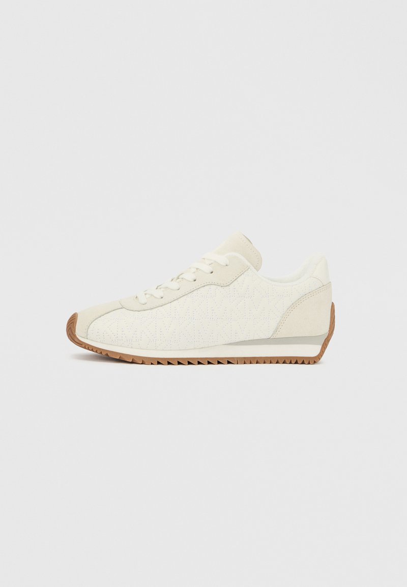 Λευκό υφασμάτινο αθλητικό παπούτσι με εξωτερική σόλα από καουτσούκ καφέ χρώματος, με χαμηλό προφίλ, στρογγυλή μύτη και κλείσιμο με κορδόνια. Λεπτομέρειες στο πλάι περιλαμβάνουν ένα μοτίβο.