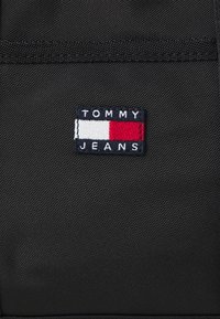 Musta kangas kott, millel on ristkülikukujuline sinine plekk, millel on valge kiri "TOMMY" ja mustal "JEANS" valgetes tähtedes, punase ja valge horisontaalse ribadisainiga.