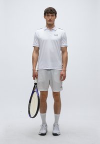 Biele polo s krátkym rukávom a jemným logom, v kombinácii s rovnakými šortkami. Drží tenisovú raketu, má na nohách biele športové topánky a ponožky.