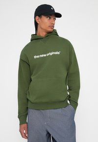 Olivno zeleno kapuco z kenguru žepom, ki ima belo besedilo "novi originali" čez prsa. Mehak, bombažen material, sproščen kroj.