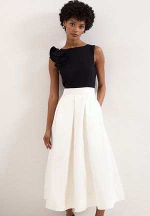 Femme portant un top noir sans manches avec un détail à volants et une jupe midi blanche taille haute, debout devant un fond clair uni.