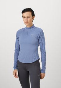 Langermet blå sportsgenser med halv glidelås, med prikkete tekstur på ermene og avrundet søm, stylet med mørke leggings.