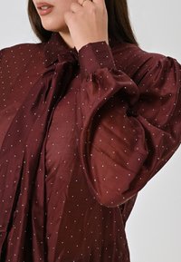 Burgunderfarbene Bluse mit Schleifenkragen, strukturiertem Stoff und weißen Punkten. Mit geschäfteten Ärmeln und einem lässigen, fließenden Design.