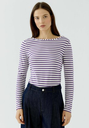 Femme aux cheveux bruns lisses porte une chemise blanche à manches longues avec des rayures horizontales violettes et un jean taille haute foncé, debout devant un fond blanc.