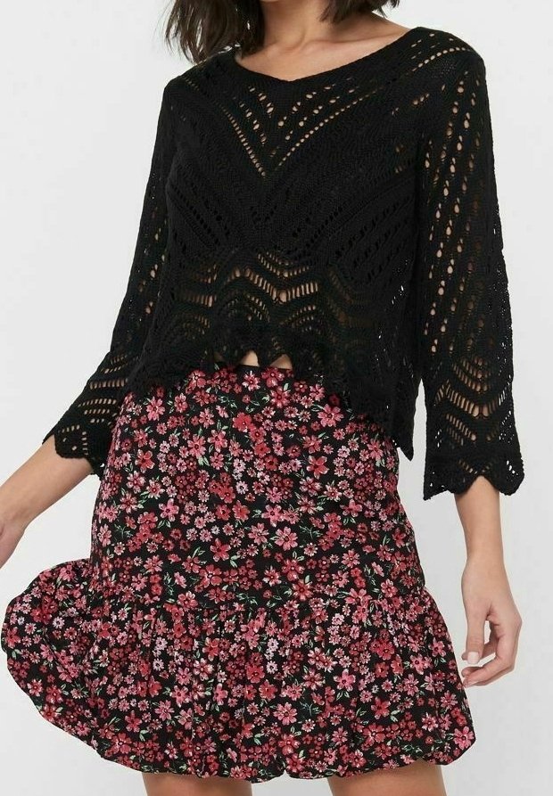 Schwarzes gestricktes Crop-Top mit offenen Mustern und gewellten Kanten, kombiniert mit einem schwarzen floralen Rüschensrock mit rosa und grünen Akzenten.