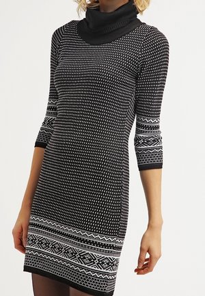 Vrouw draagt een zwart-witte gebreide jurk met driekwartsmouwen, een coltrui en geometrisch patroon op de mouwen en zoom.