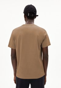 Bruine katoenen t-shirt met korte mouwen, ronde halslijn en een rechte zoom. Een zwarte pet wordt gedragen, met een verstelbare band aan de achterkant.