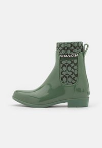 Botas de tobillo verdes de goma con acabado brillante. Parte superior de tejido de punto con un patrón verde oscuro y el logo de Coach. Tacón bajo y tira para poner.