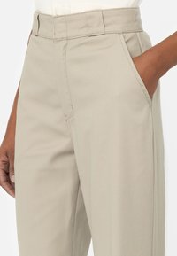 Detailní záběr na osobu v béžových kalhotách na míru, s pravou rukou v kapse na pozadí bílé barvy.