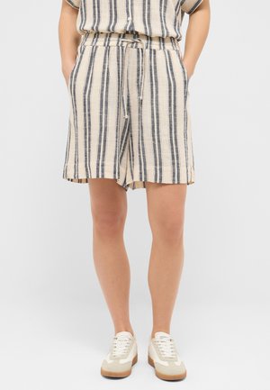 Person trägt beige und schwarze vertikal gestreifte Shorts mit Taschen und weiße Schnür-Sneaker, die vor einem schlichten Hintergrund stehen.