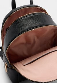 Schwarzer Texturrucksack mit goldenen Reißverschlüssen. Das Innenfutter hat eine braune Beschichtung und ein pinkes Fach mit Klappenverschluss. Mehrere Reißverschlussfächer.