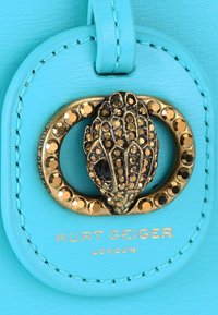 Błękitna skórzana metka ma złoty okrągły element z teksturowanym czarnym wzorem ozdobionym rhinestonami. Napis "KURT GEIGER LONDON" jest wytłoczony.