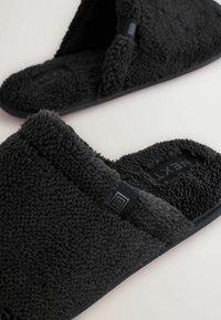 Svarta fluffiga tofflor med ett mjukt, texturerat tyg och en öppen tådesign. Har en platt sula och en liten logotyp på sidan.