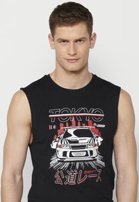 Must beebi, millel on graafiline trükk autost, mille kohal on tekst "TOKYO" ja "DRIFT" punases ja valges värvitoonis, linnavaatega taustal.