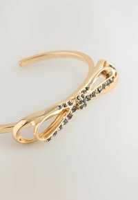 Goldfarbener Metallarmreif mit einer Schleifenform, akzentuiert mit kleinen dunklen Edelsteinen entlang der Mitte. Glatte Textur und schlichtes Design.