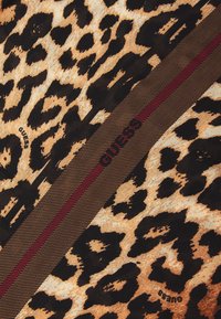 Tessuto in stampa leopardata con macchie beige e nere, accentuato da una banda diagonale rossa e dal logo "GUESS" in nero.