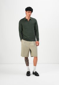 Vyras su juodais plaukais, vilkintis tamsiai žalią ilgomis rankovėmis zip-up marškinėlį, smėlio spalvos šortus, baltas kojines, juodąsias loafer'ius ir tatuiruotę ant kairiojo blauzdos.