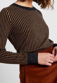 Zwart en bruin gestreepte gebreide trui met metallic draaddetails, ronde halslijn en lange mouwen, gecombineerd met een oranje corduroy rok.