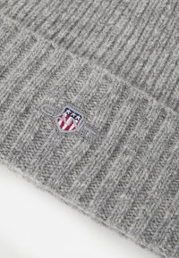 Grå ribbet lue med et teksturert strikkedesign. Har et lite brodert emblem med røde, hvite og blå elementer.