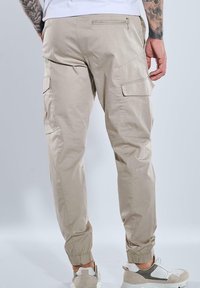Beige Cargo-Hosen mit Seiten- und Gesäßtaschen, elastischen Bündchen und einer glatten Textur. Getragen mit hellen Sneakers.