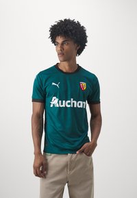 Turkos fotbollströja med korta ärmar, svart kant och mönstrade detaljer. Logotyp på bröstet och stor vit text "Auchan" som är tydligt synlig.